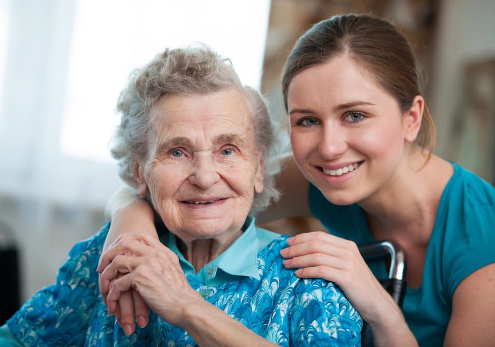 Uśmiechnięta seniorka z opiekunką, która obejmuje ją w domu